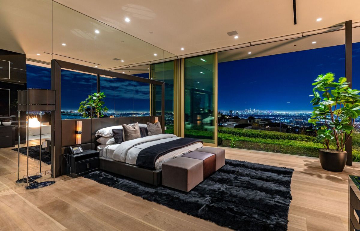 The space is a wonderful fusion of contemporary aesthetics, glamour, and mid-century rustic. Soft accents, such as airy glass doors and plush fur rugs, balance out the raw wood headboard. On either side of the bed are matching lamps. An interesting contrast to the other furniture is the abstract wall on the right.