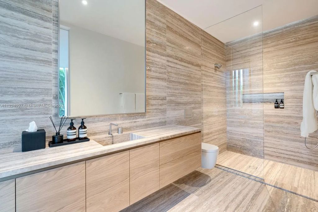 28 W Di Lido Drive, Miami Beach, Florida is stunning home where relaxed island living meets worldly sophistication, incredible features include 12ft ceilings, column-free living, 54ft of automated glass, media bookshelf, wine bar, & vapor fireplace highlight the textured travertine and white oak palette.