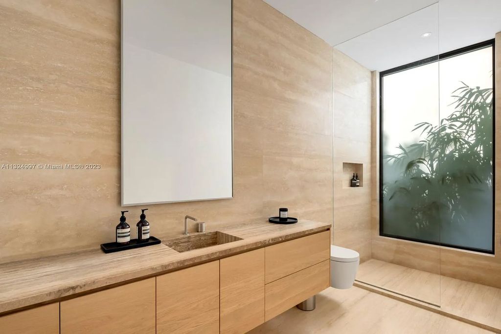 28 W Di Lido Drive, Miami Beach, Florida is stunning home where relaxed island living meets worldly sophistication, incredible features include 12ft ceilings, column-free living, 54ft of automated glass, media bookshelf, wine bar, & vapor fireplace highlight the textured travertine and white oak palette.