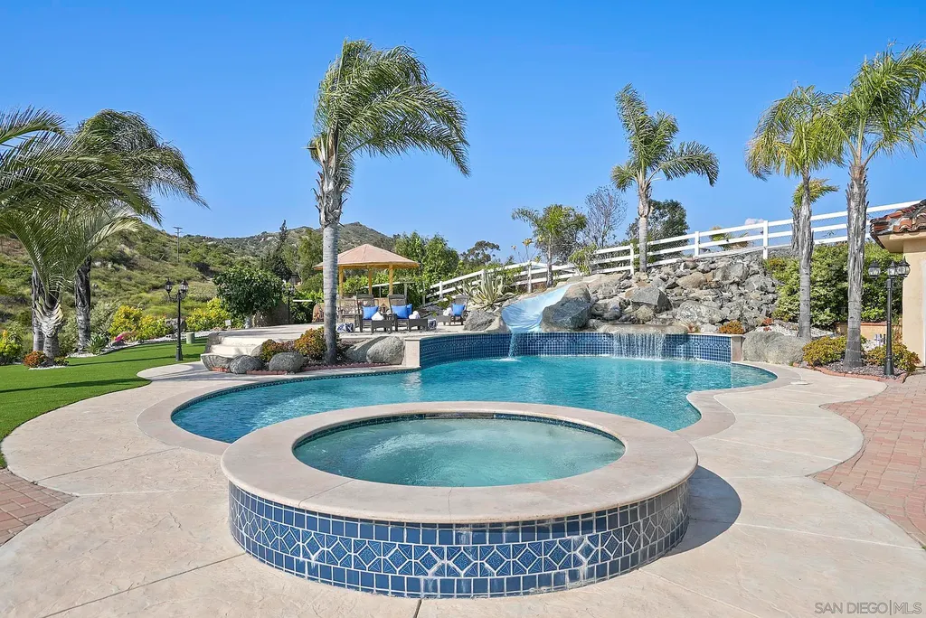 Elevated on a hill in Blossom Valley, this exquisite home offers a living experience that is truly unmatched. Nestled within a gated estate, the property expands across six acres of meticulously landscaped grounds, showcasing natural rock formations and a vibrant variety of palm trees, succulents, and blooming flowers. Embracing eco-friendly features such as a ground-water well and solar panels, this residence seamlessly blends sustainability with luxurious living. 15987 Broad Oaks Road Home in El Cajon, California.