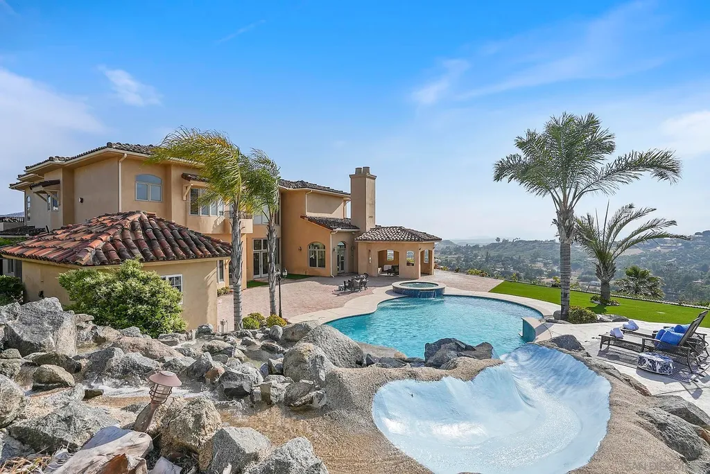 Elevated on a hill in Blossom Valley, this exquisite home offers a living experience that is truly unmatched. Nestled within a gated estate, the property expands across six acres of meticulously landscaped grounds, showcasing natural rock formations and a vibrant variety of palm trees, succulents, and blooming flowers. Embracing eco-friendly features such as a ground-water well and solar panels, this residence seamlessly blends sustainability with luxurious living. 15987 Broad Oaks Road Home in El Cajon, California.