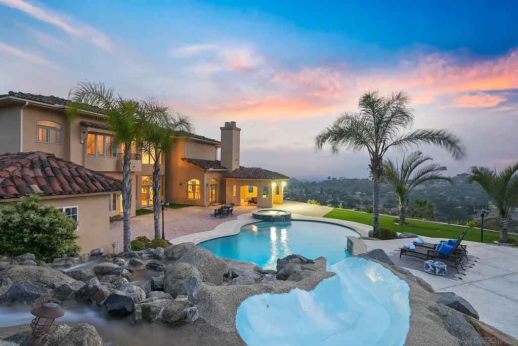 Elevated on a hill in Blossom Valley, this exquisite home offers a living experience that is truly unmatched. Nestled within a gated estate, the property expands across six acres of meticulously landscaped grounds, showcasing natural rock formations and a vibrant variety of palm trees, succulents, and blooming flowers. Embracing eco-friendly features such as a ground-water well and solar panels, this residence seamlessly blends sustainability with luxurious living. 15987 Broad Oaks Road Home in El Cajon, California.