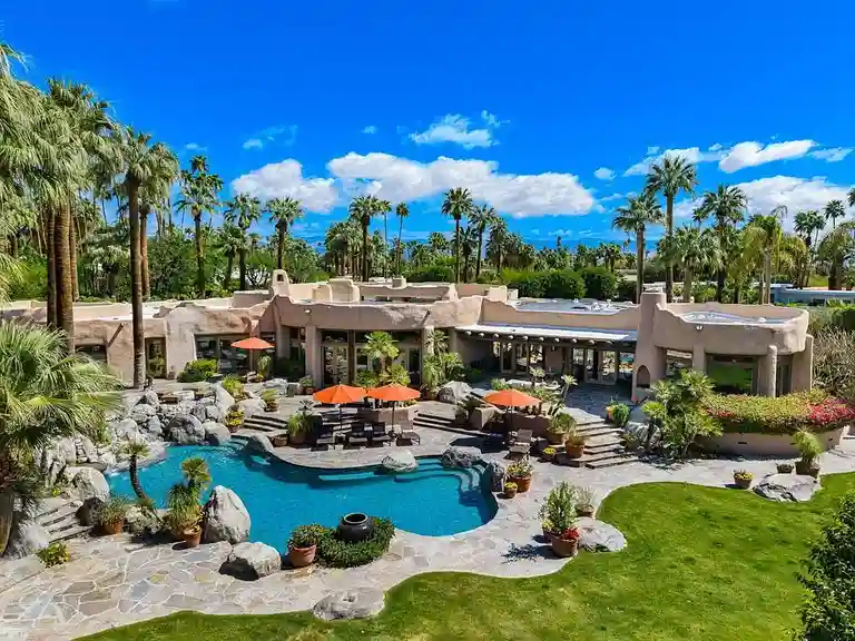Aerial view of the resort-style pool area and the main residence at 475 S Via Las Palmas, surrounded by lush palm tree groves and offering stunning mountain backdrop, highlighting the expansive 1.88-acre lot of the Most Expensive Home in Palm Springs.