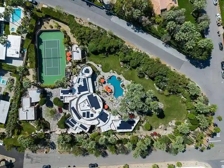 High aerial view of the 475 S Via Las Palmas compound showing the main residence, resort pool, full-size tennis court, and the substantial solar array on the roof, highlighting the 1.88-acre lot size.