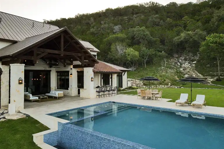 The expansive pool area and covered patio of a **luxury home in Austin**, featuring a rectangular pool with mosaic tiles, an outdoor bar, dining area, and lush green hillside backdrop, highlighting an **expensive home in Austin**.