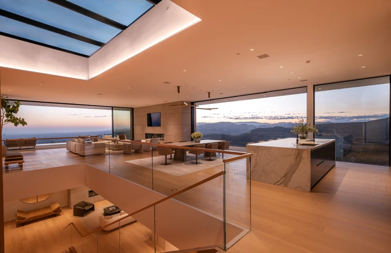 Open-plan great room with a skylight, marble kitchen island, dining area, and full-width glass walls showcasing the view.