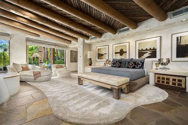 The primary bedroom of this luxury home Palm Springs, featuring a king-size bed, elegant furnishings, flagstone flooring, and large windows looking out to the lush gardens, within the 475 S Via Las Palmas estate.