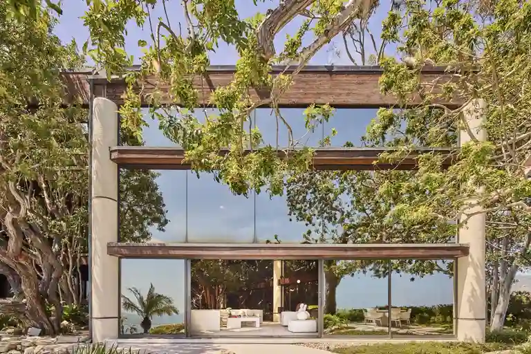 The ocean-facing facade of the estate, featuring massive floor-to-ceiling glass walls reflecting trees and the sky, highlighting the modernist design.
