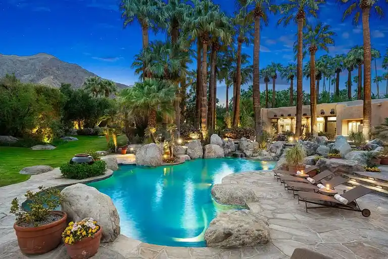The luxurious resort-style pool at 475 S Via Las Palmas illuminated at dusk, with ambient lighting, lounge chairs, and the San Jacinto Mountains in the background, making it a standout feature of this home in Palm Springs for sale.