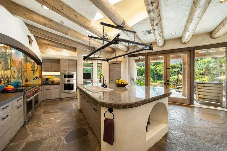 The newly reimagined chef's kitchen featuring a large granite island, professional Wolf range, integrated appliances (Miele/SubZero), rustic log ceiling beams, and a bold, desert-themed tile backsplash.