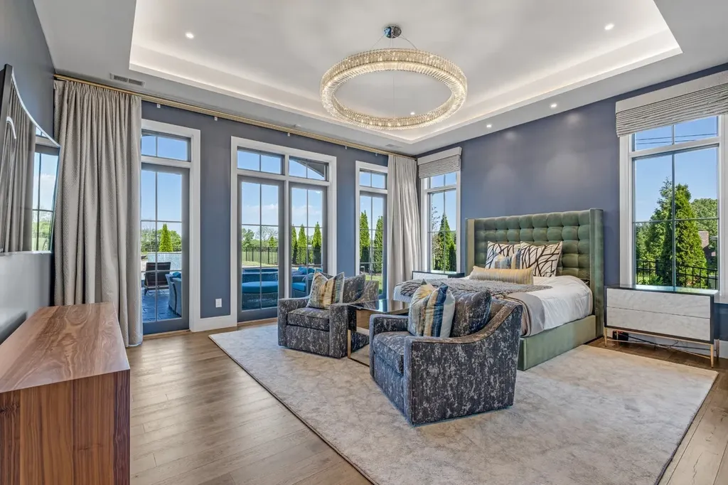 Primary bedroom with floor-to-ceiling windows, modern chandelier, tufted green headboard, and seating area in a luxury home in Mount Juliet, Tennessee.