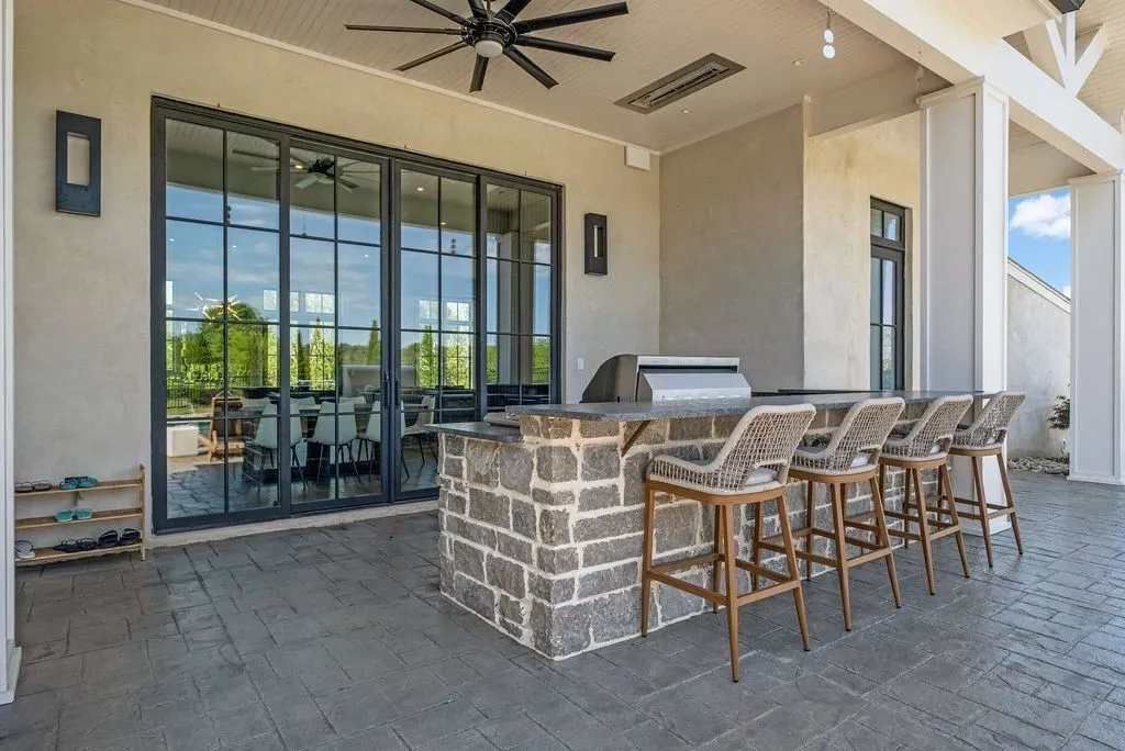 Outdoor kitchen and bar with stone island, rattan bar stools, and sliding glass doors at a luxury home in Mount Juliet, Tennessee.