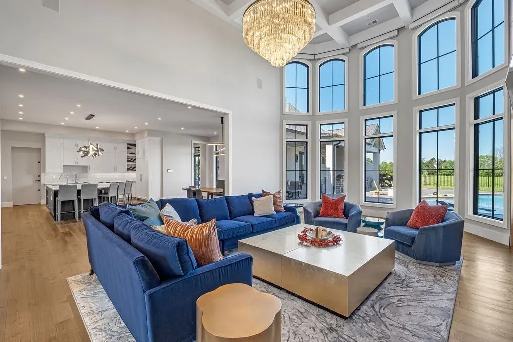 Open-concept living room with floor-to-ceiling windows, blue seating, chandelier, and modern kitchen view in a Mount Juliet luxury home.