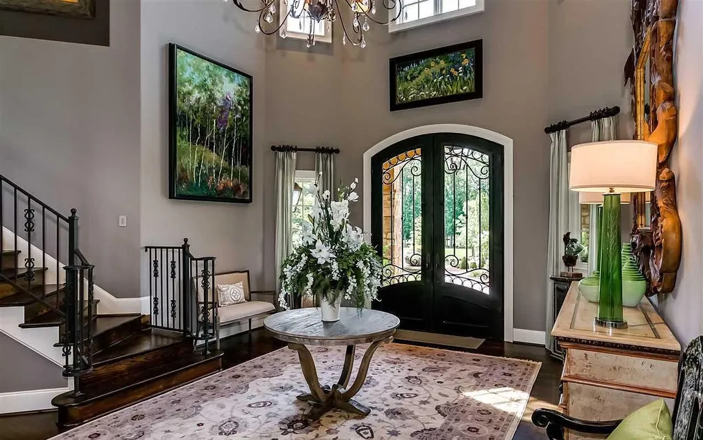 Elegant foyer of the luxury home at 2470 Cahaba Valley Rd, Indian Springs, Alabama, featuring arched wrought-iron double doors, dark hardwood stairs, fine art, and floral decor, as featured in Luxury Houses Magazine.