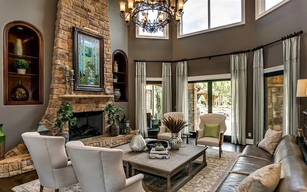 Grand living room at 2470 Cahaba Valley Rd, Indian Springs, Alabama, featuring a soaring stone fireplace, elegant chandelier, tall windows, and classic neutral interiors, highlighted in Luxury Houses Magazine.