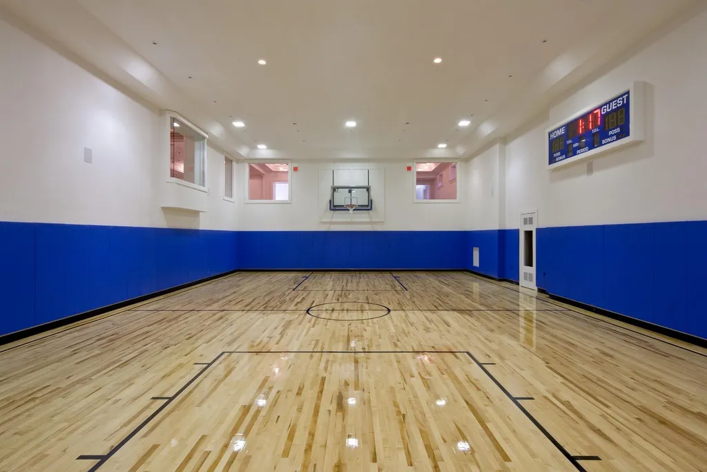 Step Inside One of America’s Most Extraordinary Modern Estates in Alpine 10 Indoor basketball court at 1 Frick Drive Alpine New Jersey featuring polished hardwood floors blue wall padding central hoop and electronic scoreboard.