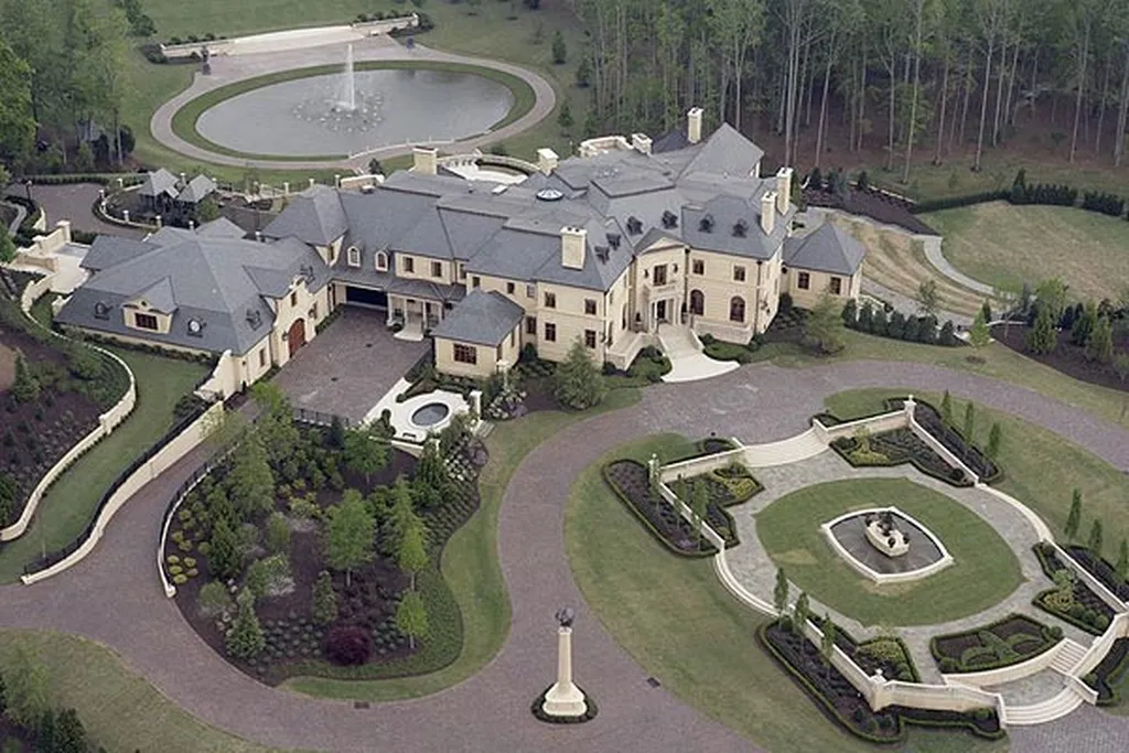 An aerial view of the luxurious Le Rêve estate in Cumming, Georgia. The mansion has a cream-colored facade and a dark gray roof, situated amid extensive manicured grounds. Features include a large circular front garden with ornate hedges, a massive winding driveway, and a large decorative pond with a fountain in the background