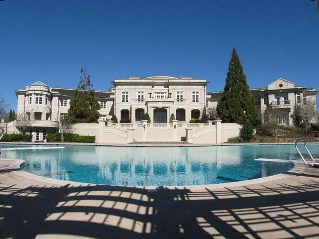 A grand, symmetrical view of the white facade of The Promise Land mansion as seen across the large, curved main swimming pool. The pool area features a diving board and surrounding patio. The mansion's central section includes a recessed portico and two sweeping staircases leading up from the pool level.