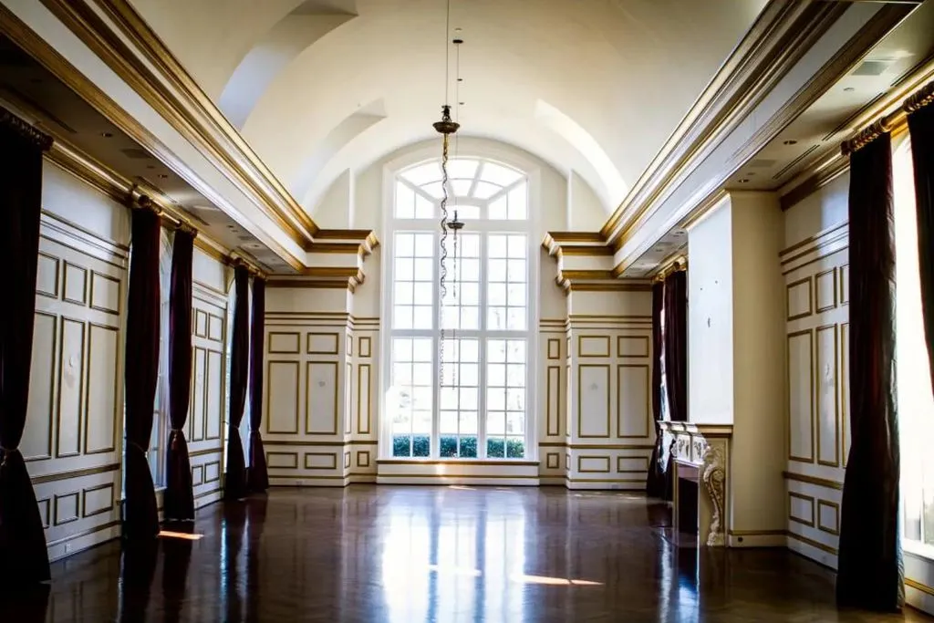 A wide, empty formal hall inside The Promise Land mansion. The room features a vaulted ceiling, polished dark hardwood floors, and walls adorned with white paneling outlined in luxurious gold trim. A massive arched window at the far end and long burgundy drapes flanking the side windows emphasize the room's grandeur.