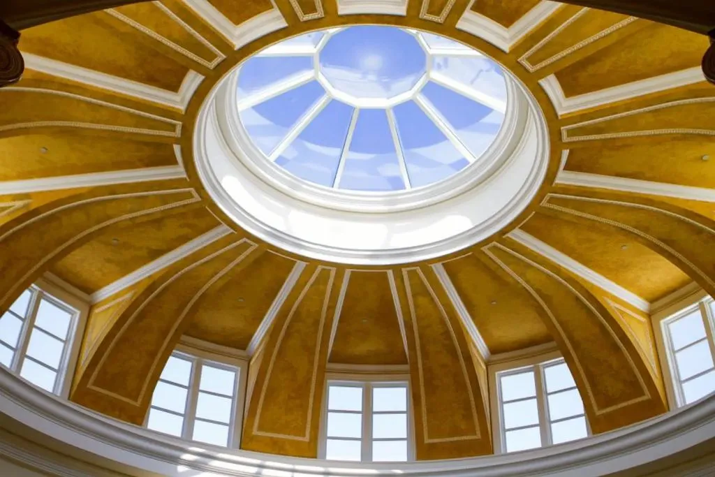An upward-facing close-up view of the interior dome of The Promise Land mansion's rotunda. The dome is painted in a rich, textured gold color with white trim and features a large central circular skylight (oculus) showcasing the bright blue sky above