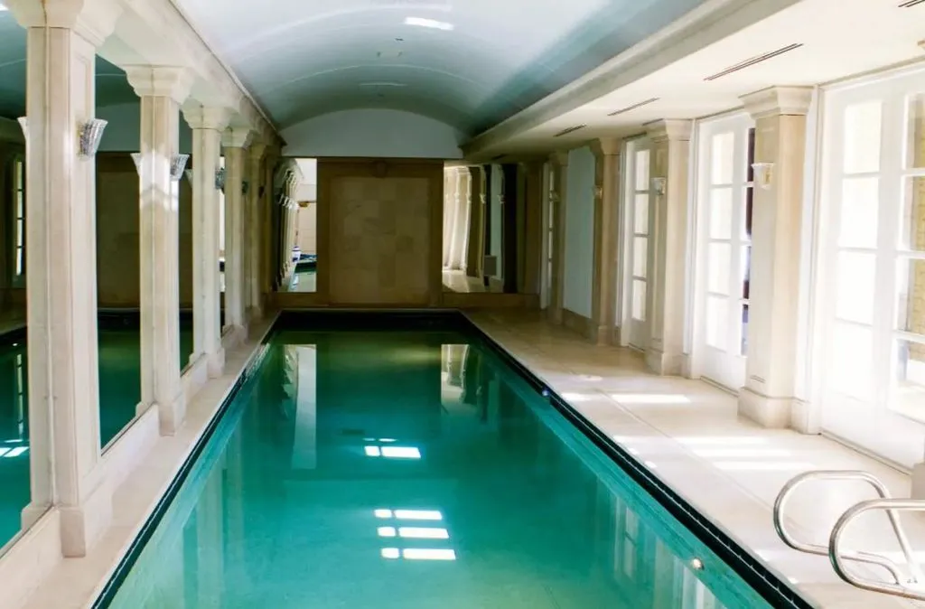 A long, narrow indoor lap pool inside The Promise Land mansion. The pool is lined on one side by a colonnade of white Roman-style pillars, and on the other by tall French doors leading to the exterior. The ceiling is a vaulted arch, and the entire space is finished in light marble or stone, giving it a classic, elegant spa feel