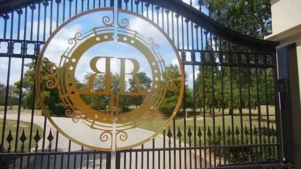 A close-up view of the custom-made wrought iron entrance gates of The Promise Land. The center features a large, reflective gold circular emblem with the intertwined letters 'RR', representing Rick Ross, set against the backdrop of the long driveway and surrounding greenery.