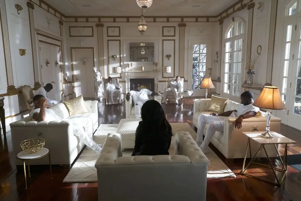 A luxuriously furnished living room inside The Promise Land mansion, featuring white couches and chairs, set against walls with elegant white and gold trim. Several men, including Rick Ross, and a woman are lounging in the room, all wearing white or dark clothing. The room boasts a marble fireplace, French doors, and crystal chandeliers.