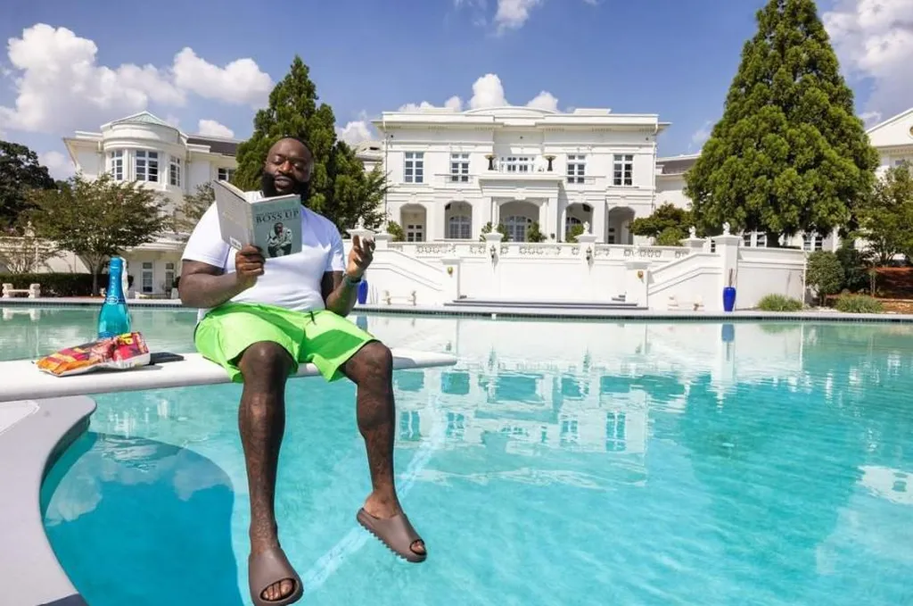 Rapper Rick Ross is sitting on a diving board by a turquoise pool, reading his book "Boss Up" in front of his white Promise Land mansion under the bright sun. A large water bottle and snacks are visible next to him