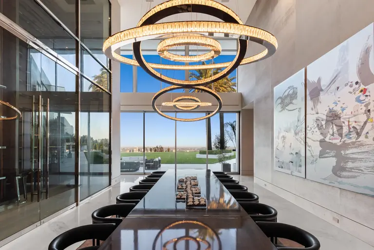 The elegant formal dining room inside The One, featuring a high ceiling, custom circular chandeliers, and panoramic views of the Los Angeles landscape.