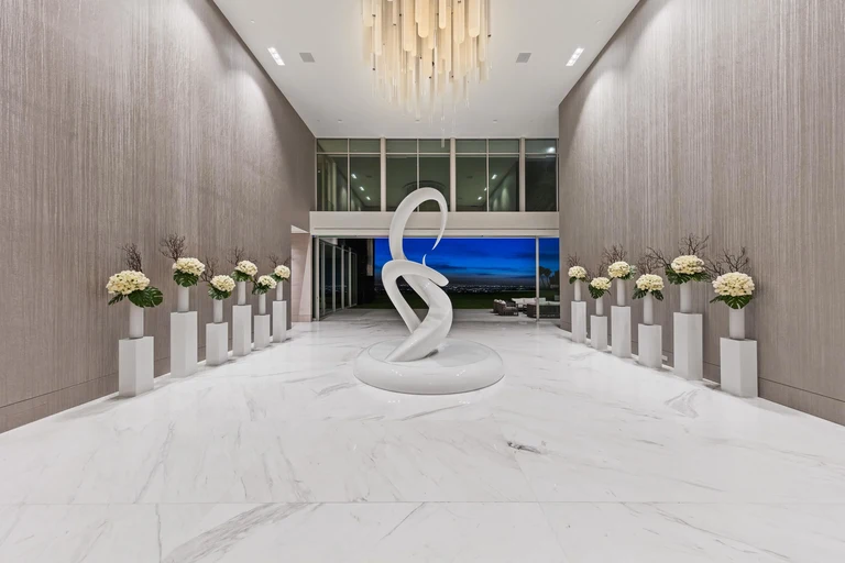 The massive, grand foyer of The One, the biggest home in Los Angeles, featuring white marble floors, a sculptural centerpiece, and floor-to-ceiling windows.