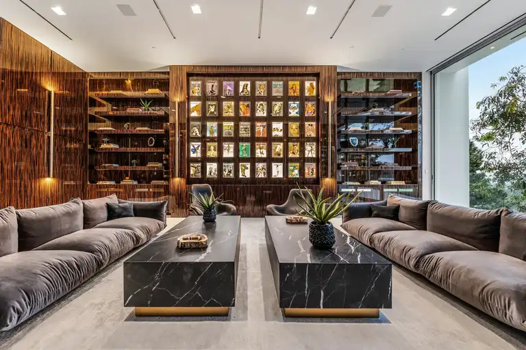 The sophisticated cigar lounge and library area inside The One, the biggest home in Los Angeles, featuring plush velvet sofas, marble tables, and backlit display shelving.