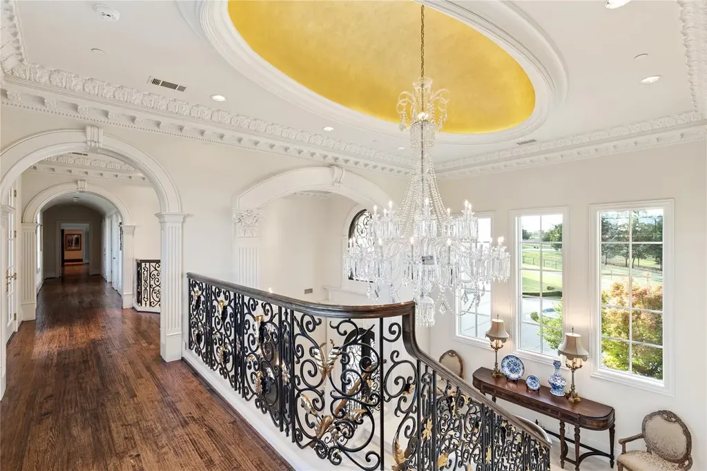 Château Lumièr in McKinney Lists for $14.65 Million as a French Chateau Estate Spanning Fifteen Wooded Acres 4 Upper gallery inside Château Lumièr at 1201 Gray Branch Rd in McKinney featuring ornate iron balustrades a crystal chandelier and a gold leaf ceiling detail.