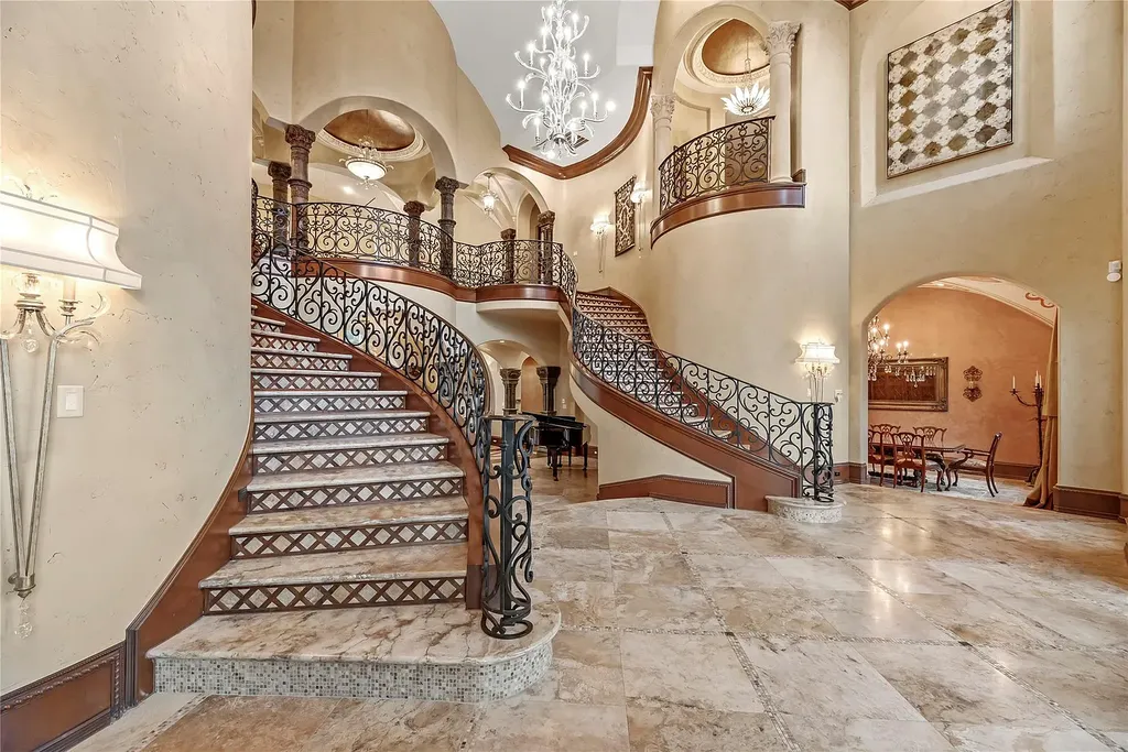 A Mediterranean Masterpiece Rising Above the Woodlands of Magnolia, Texas Seeks for $11,500,000 4 Lavish grand foyer of the Mediterranean estate at 38262 Windy Ridge Trail in Magnolia, Texas, showcasing dual curved staircases with ornate wrought-iron railings, marble flooring, and dramatic vaulted ceilings with chandeliers.