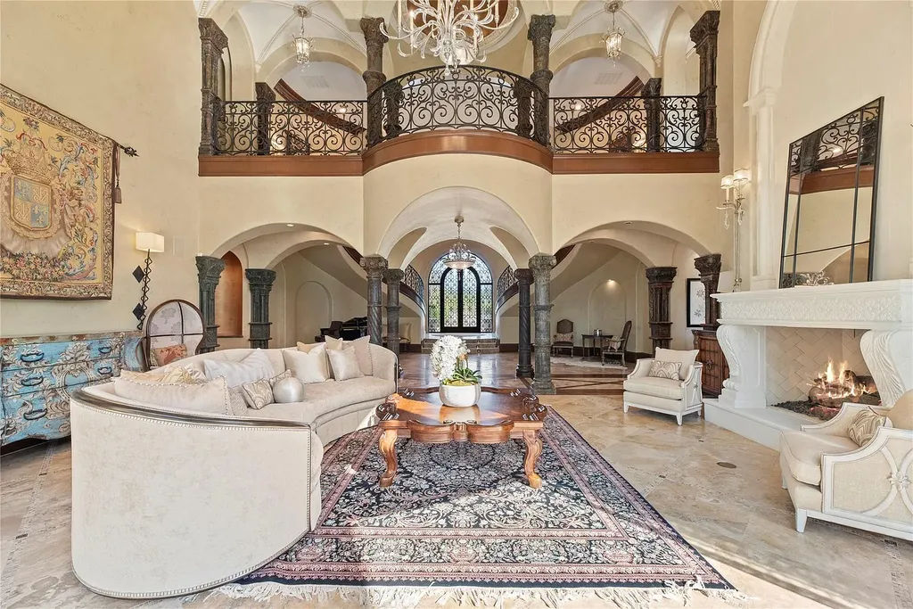 A Mediterranean Masterpiece Rising Above the Woodlands of Magnolia, Texas Seeks for $11,500,000 5 Elegant grand salon in the Mediterranean estate at 38262 Windy Ridge Trail, Magnolia, Texas, featuring soaring ceilings, wrought-iron upper galleries, stone columns, a large curved sofa, ornate rug, and a carved fireplace.