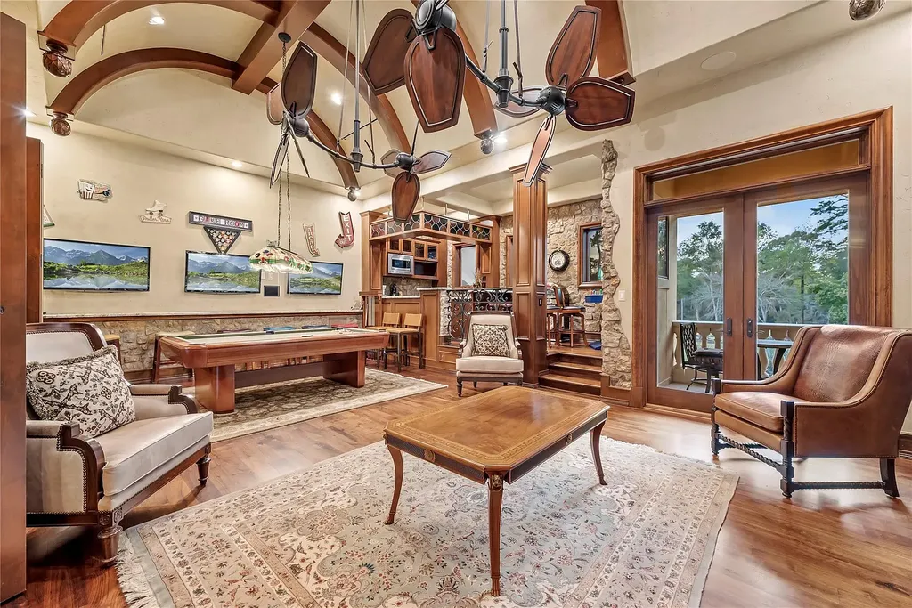 A Mediterranean Masterpiece Rising Above the Woodlands of Magnolia, Texas Seeks for $11,500,000 11 Luxury game room at 38262 Windy Ridge Trail in Magnolia, Texas, featuring a pool table, vaulted ceilings with sculptural fans, custom woodwork, stone accents, lounge seating, and doors opening to a balcony with wooded views.