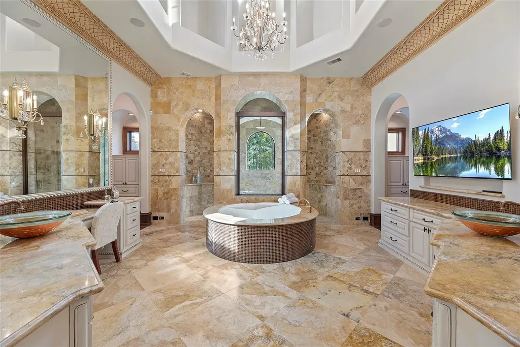 A Mediterranean Masterpiece Rising Above the Woodlands of Magnolia, Texas Seeks for $11,500,000 10 Spa-like primary bathroom at 38262 Windy Ridge Trail in Magnolia, Texas, featuring a circular soaking tub, travertine floors and walls, dual vanities, arched alcoves, chandelier lighting, and a wall-mounted TV.