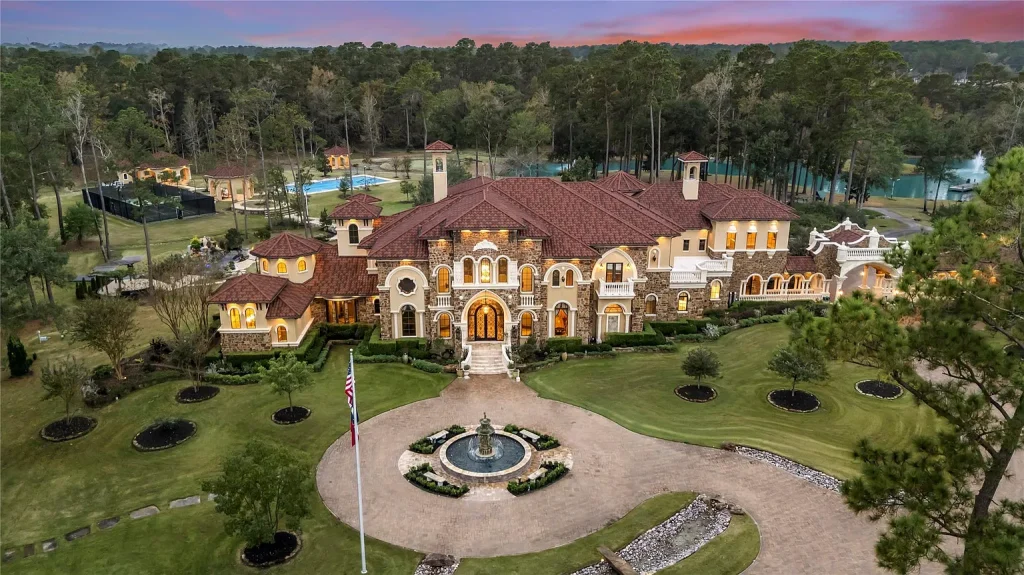 A Mediterranean Masterpiece Rising Above the Woodlands of Magnolia, Texas Seeks for $11,500,000 1 Dusk aerial view of the Mediterranean estate at 38262 Windy Ridge Trl in Magnolia, Texas, showing illuminated stone façade, red tile roof, circular driveway with fountain, landscaped grounds, tennis court, pool, and surrounding forest.
