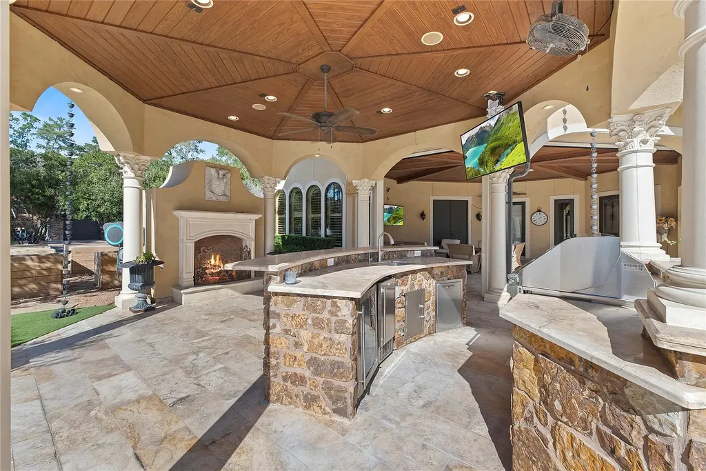 A Mediterranean Masterpiece Rising Above the Woodlands of Magnolia, Texas Seeks for $11,500,000 14 Luxurious outdoor kitchen pavilion at 38262 Windy Ridge Trail in Magnolia, Texas, featuring stone countertops, built-in grill, fireplace, vaulted wood ceiling, and columned arches overlooking the landscaped grounds.