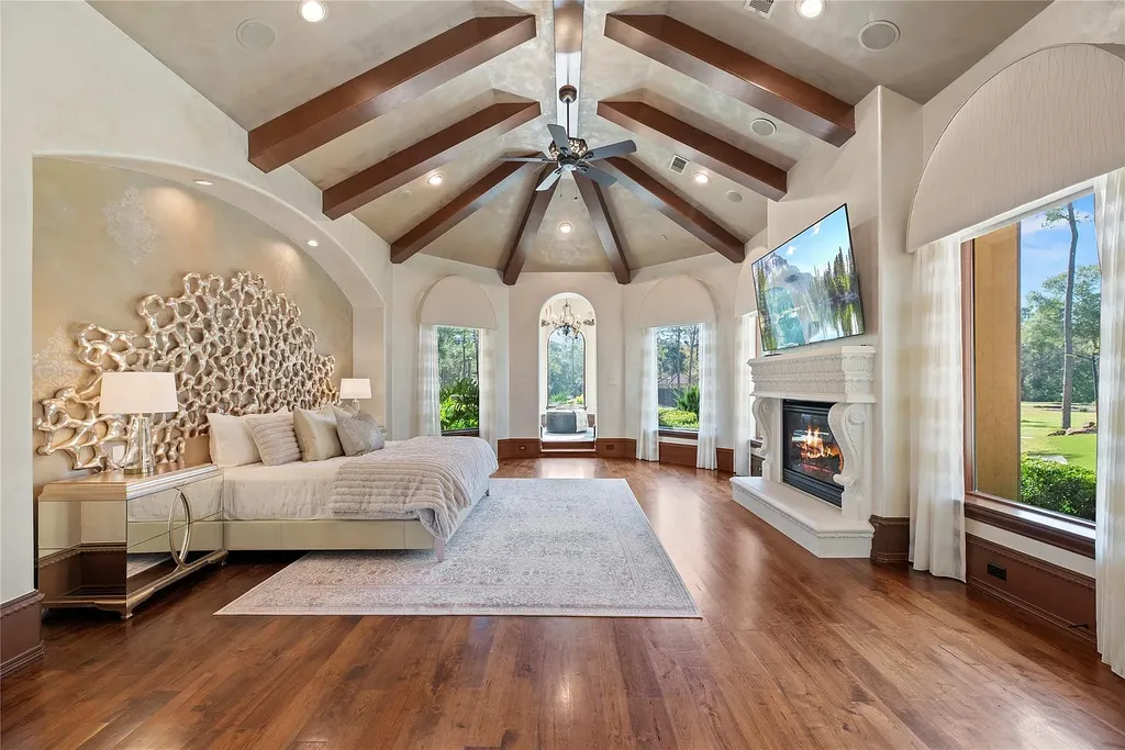 A Mediterranean Masterpiece Rising Above the Woodlands of Magnolia, Texas Seeks for $11,500,000 9 Luxurious primary bedroom at 38262 Windy Ridge Trail in Magnolia, Texas, featuring vaulted wood-beam ceilings, a sculptural headboard wall, hardwood floors, large arched windows, and an ornate fireplace with a mounted TV.